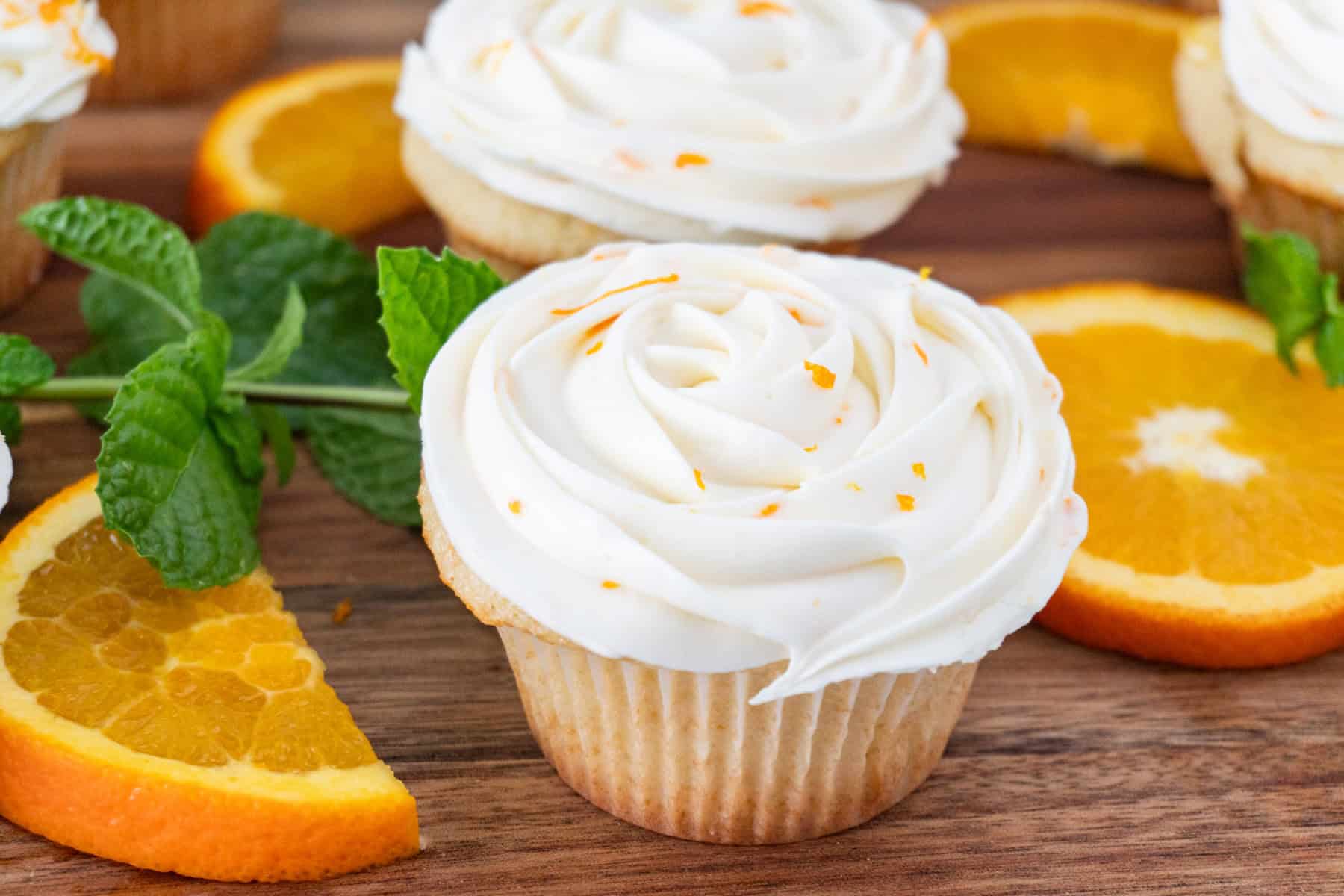 Orange cupcakes with cream cheese frosting on a wooden cutting board next to orange slices.