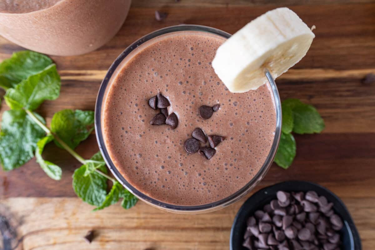 chocolate banana smoothie in glass on cutting board with banana slice and chocolate chips.