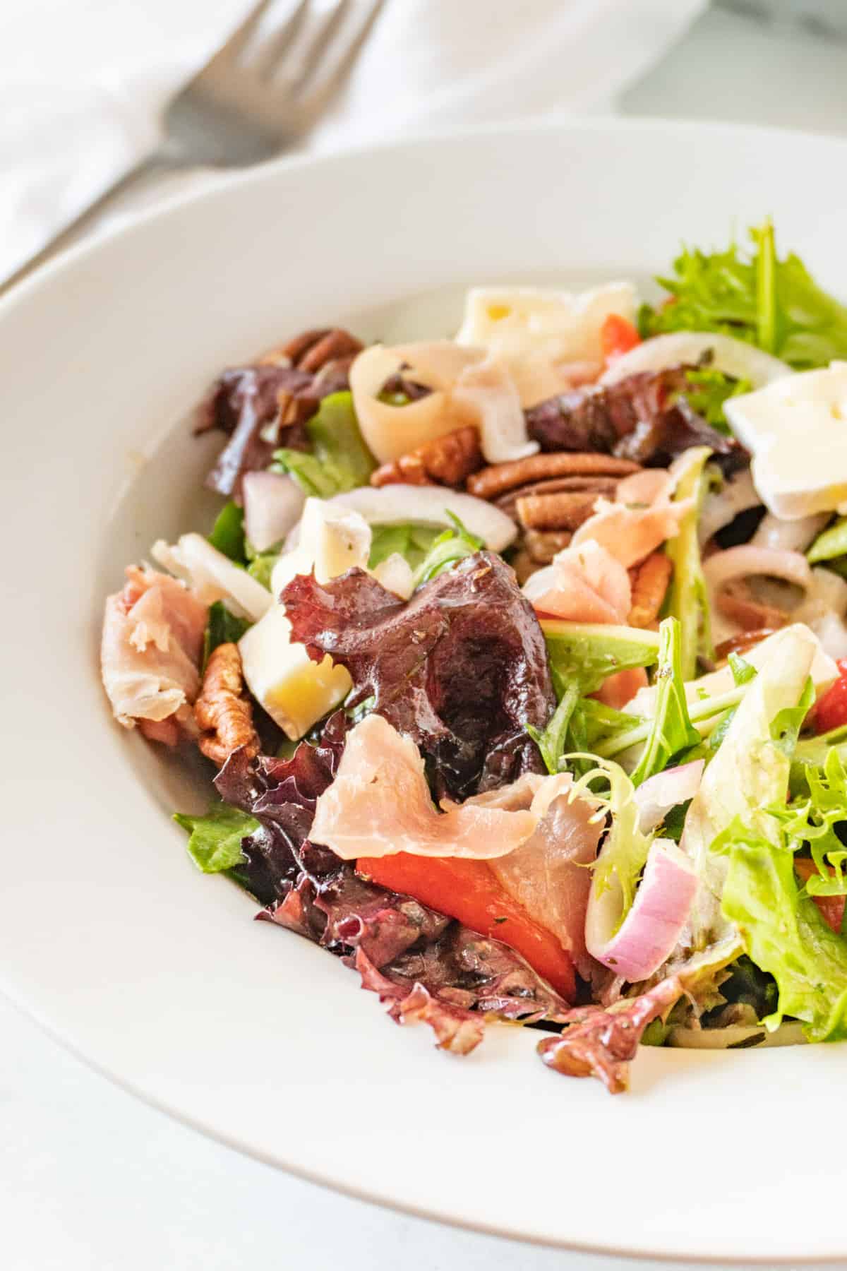 white salad bowl filled with salad mix, prosciutto, brie, red onion