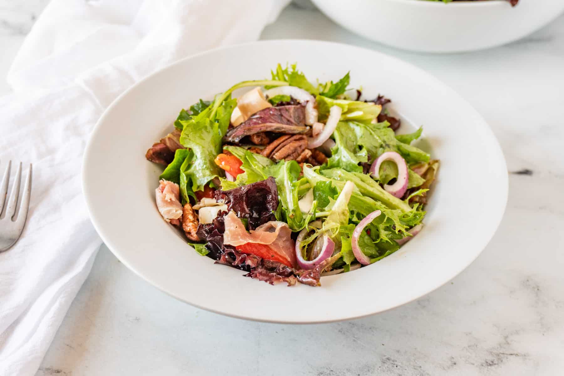 white salad bowl filled with salad mix, prosciutto, brie, red onion with fork and napkin next to it