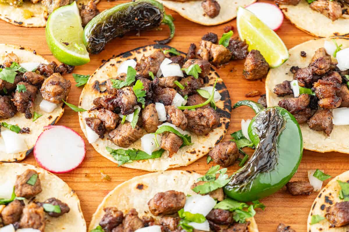 taco sprinkled with onion and cilantro on a cutting board surrounded by blackened jalapeno, radish slices and lime wedges.