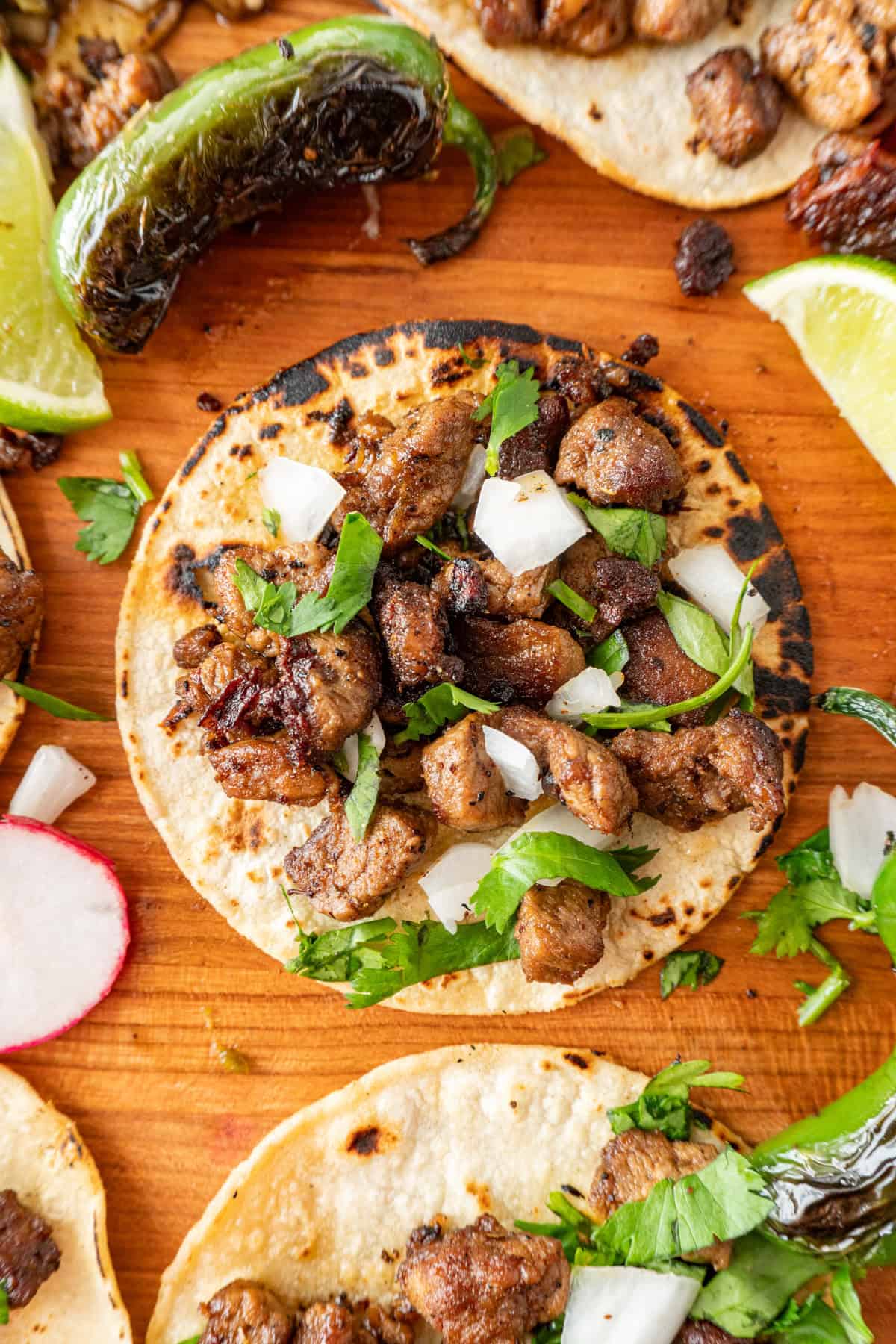 taco sprinkled with onion and cilantro on a cutting board surrounded by blackened jalapeno and lime wedges.