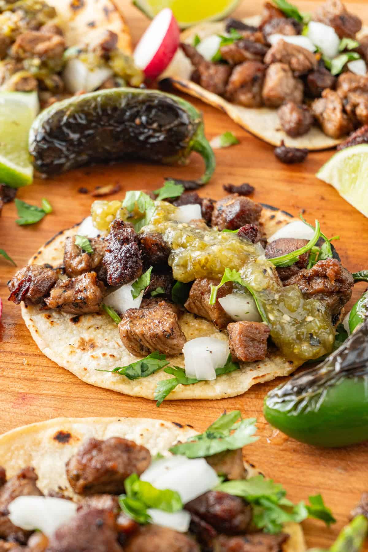 taco topped with tomatillo salsa, sprinkled with onion and cilantro on a cutting board surrounded by blackened jalapeno and lime wedges.