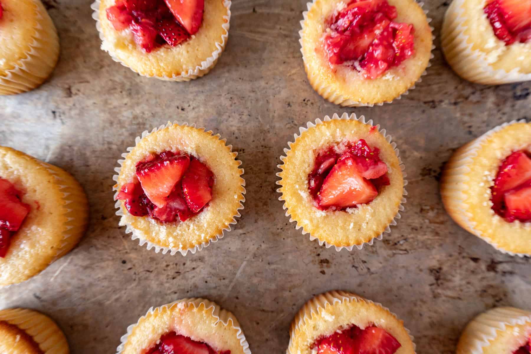 baked cupcakes on a baking sheet, with holes cut out of the center of each, and filled with strawberry filling.