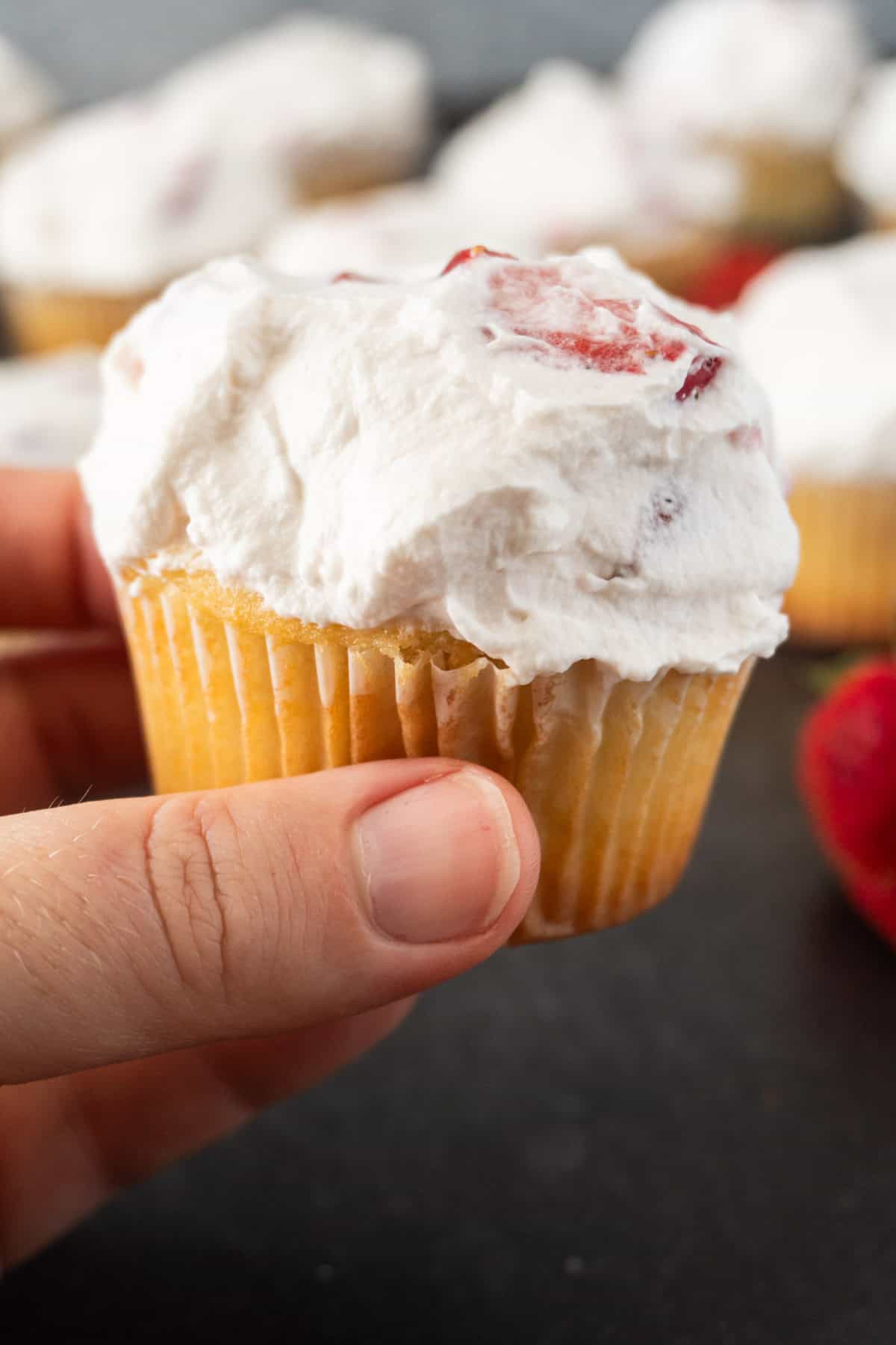 holding a strawberry shortcake cupcake, with more cupcakes in the background.