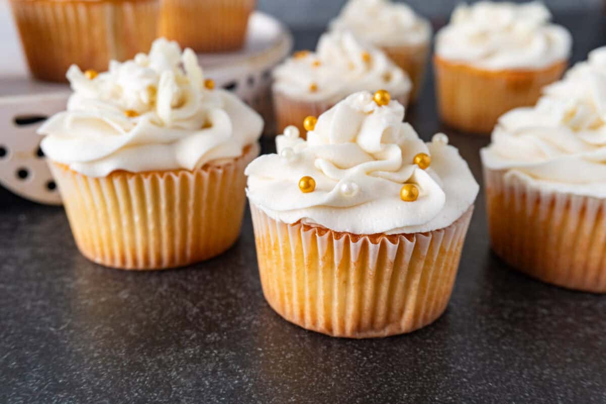 White chocolate cupcake with white chocolate frosting and gold and white sprinkles, and some with white chocolate pieces in frosting.