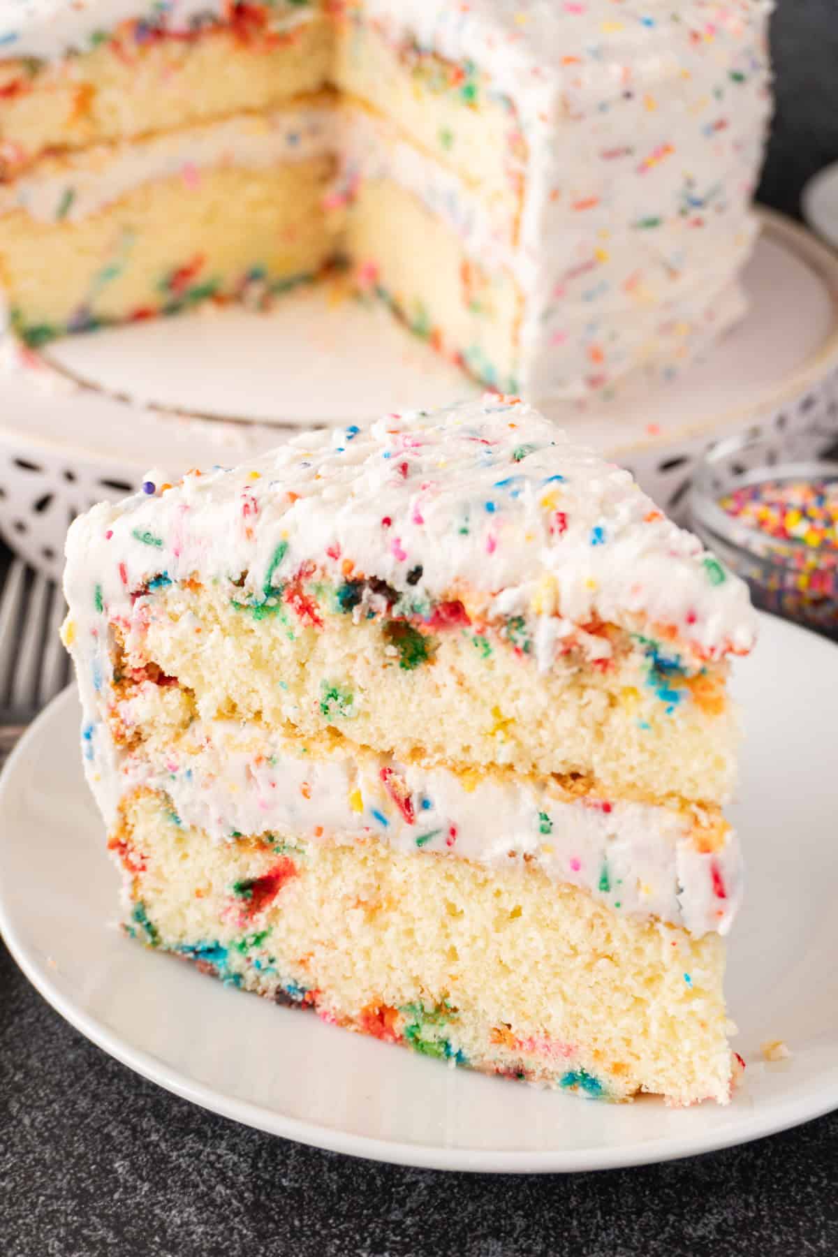 Slice of Birthday Cake on a white plate with rest of cake in background.