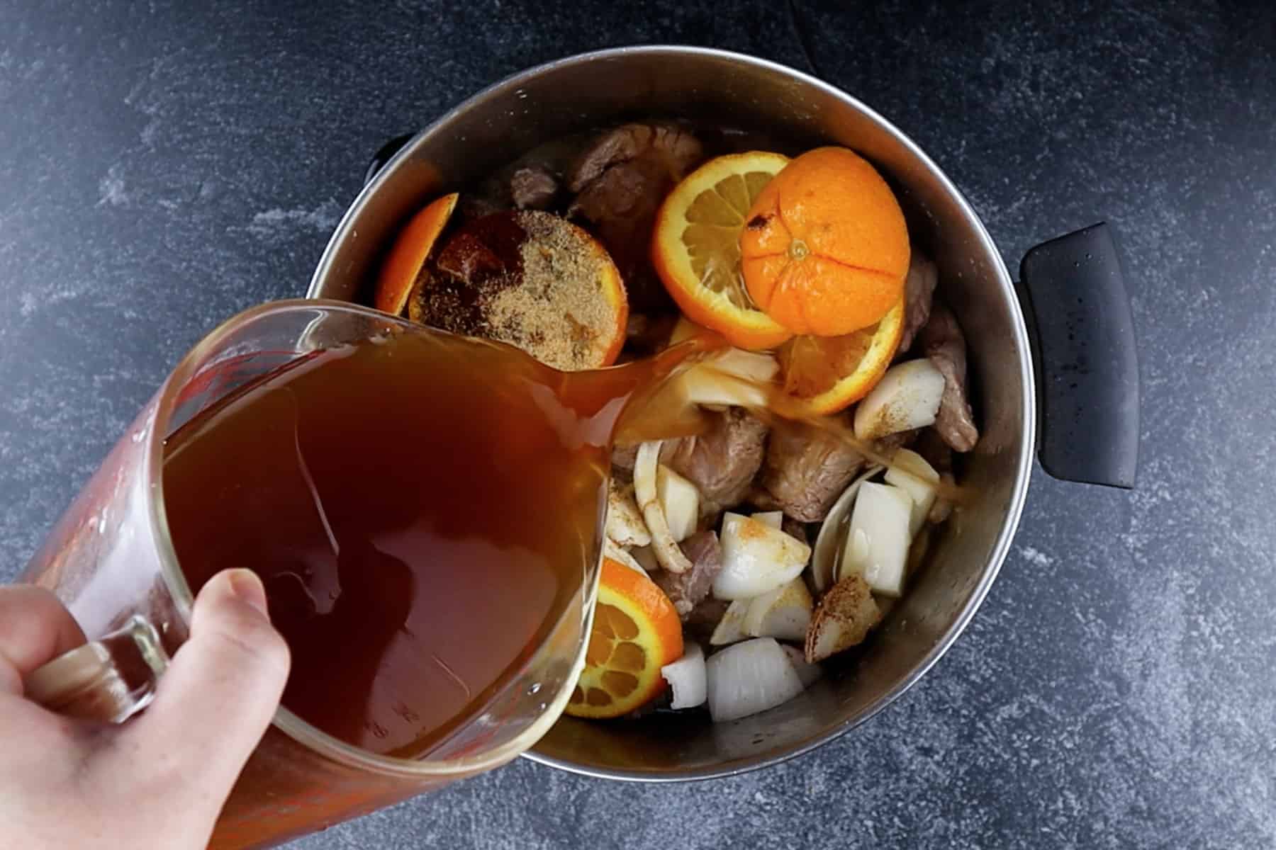Adding broth to the cooking pot with pork butt, spices, orange, and onion.