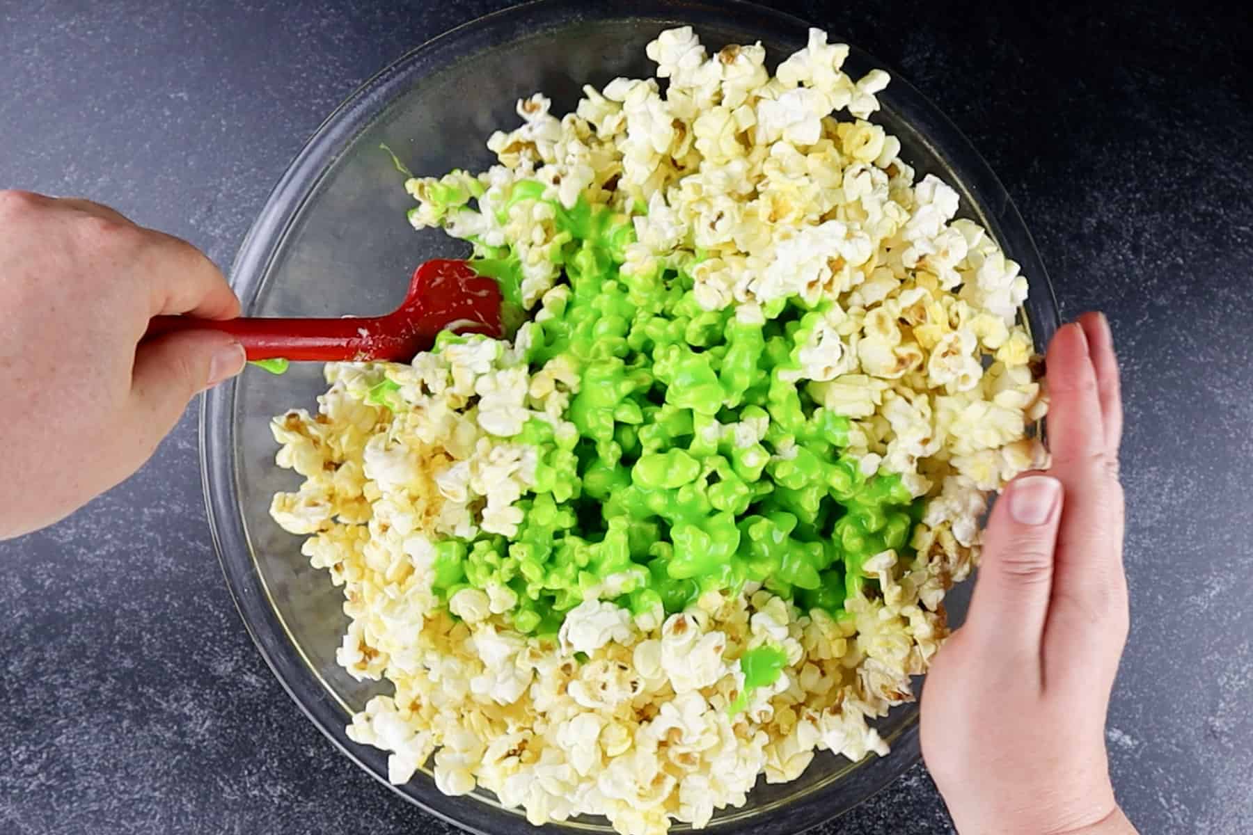 mixing the melted marshmallows into the popcorn in a clear glass bowl.