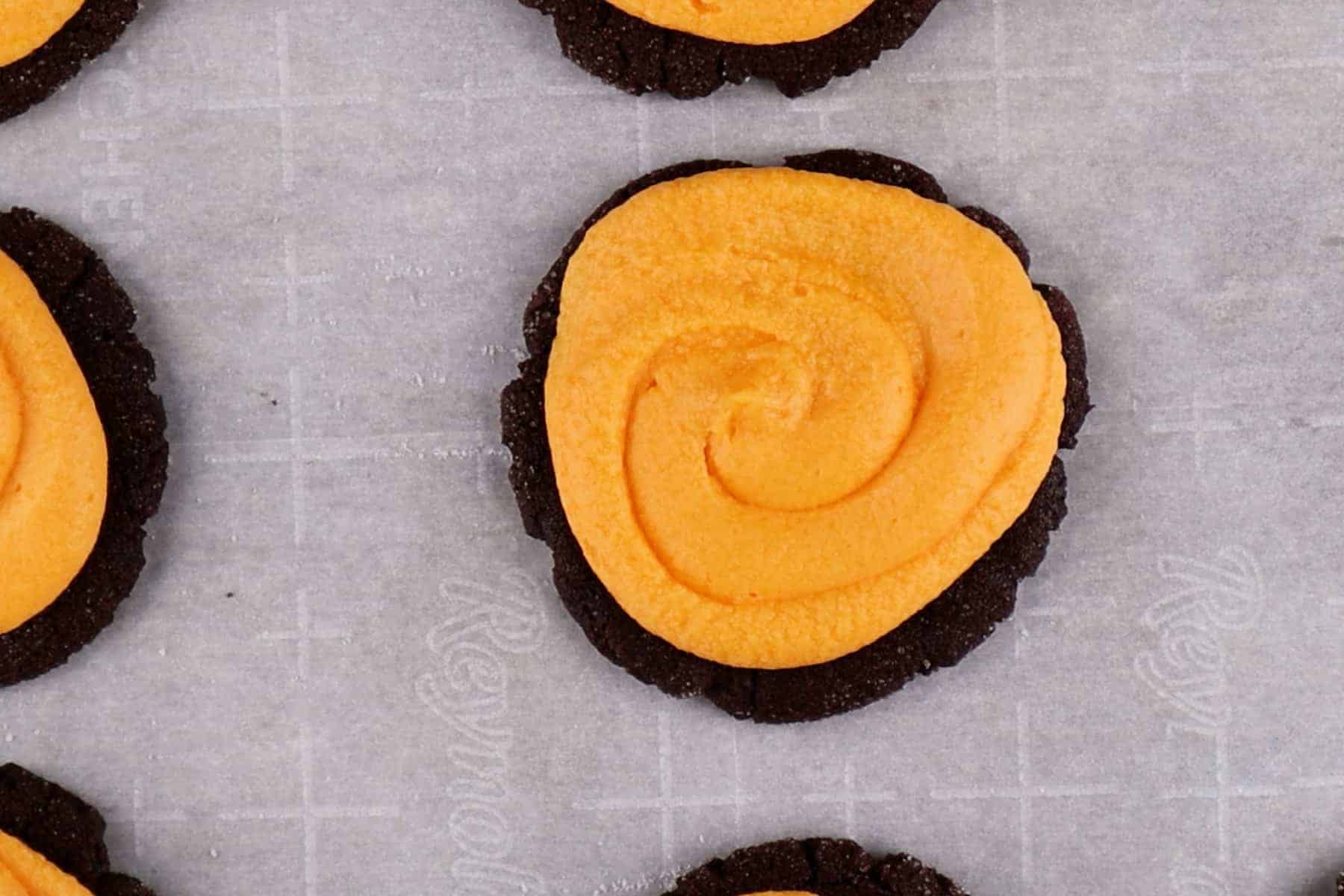 Orange colored buttercream piped on top of a chocolate sugar cookie.