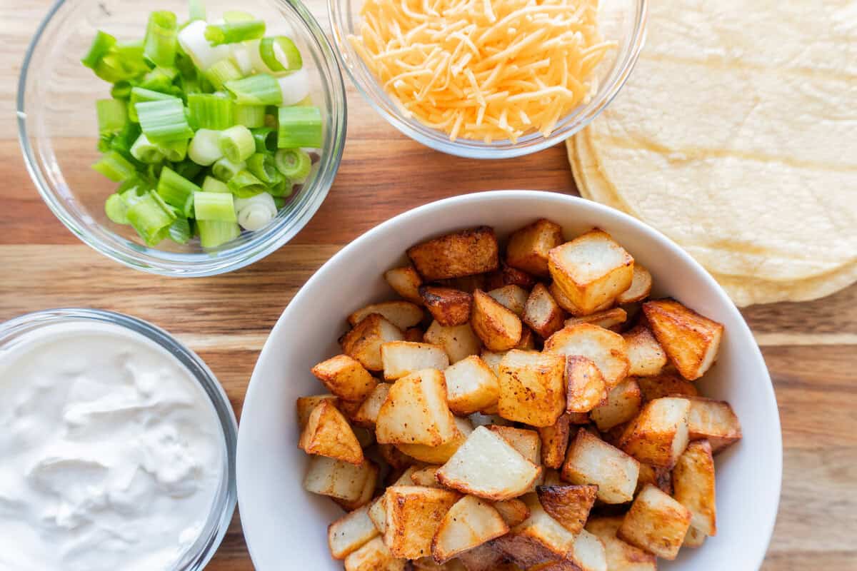 cooked potatoes in a white bowl, cheese, green onion sour cream, and tortillas next to the potatoes, on a wooden cutting board