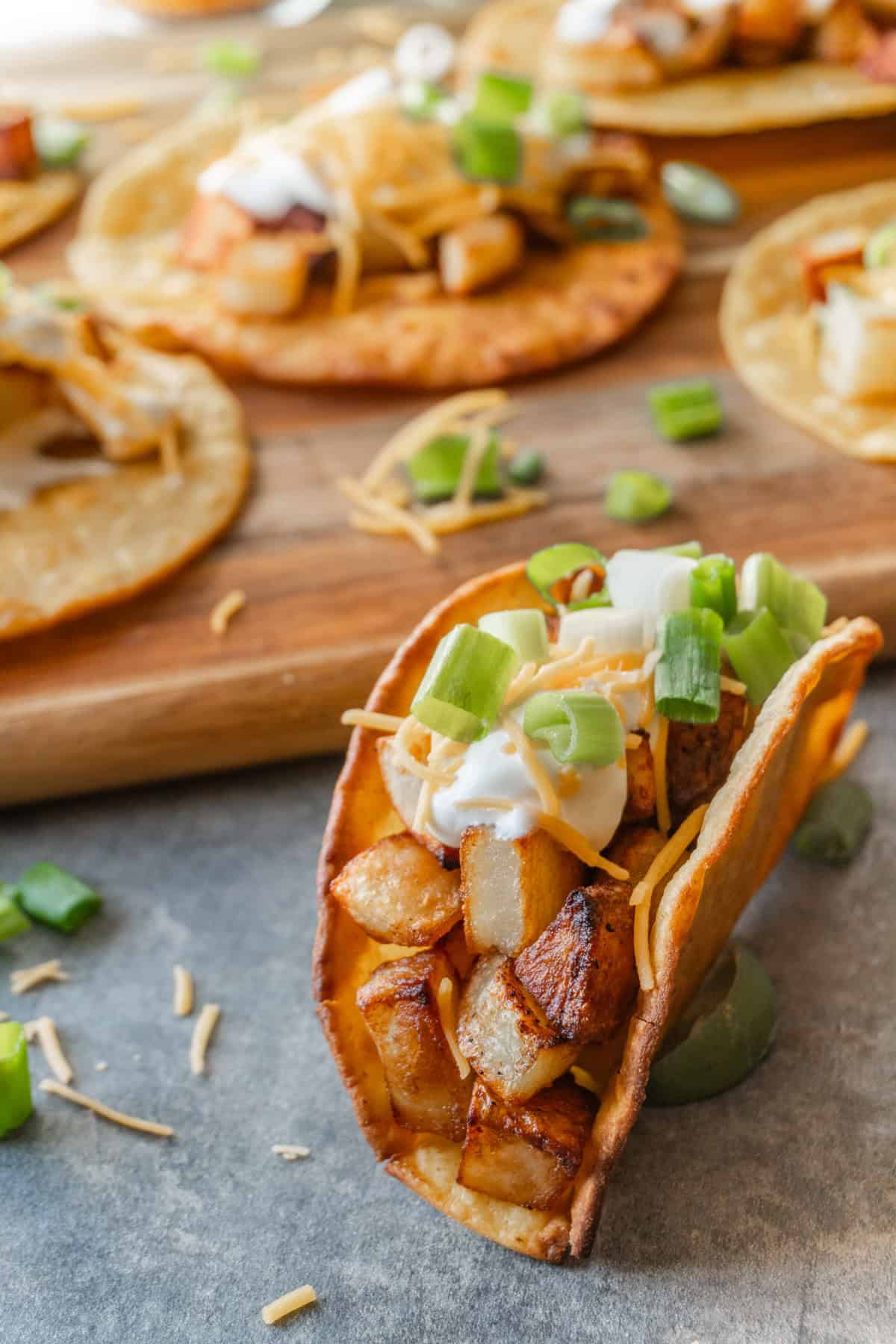 Potato taco in crispy corn tortilla topped with sour cream, cheese, and green onion with more tacos on a wooden cutting board in the background.