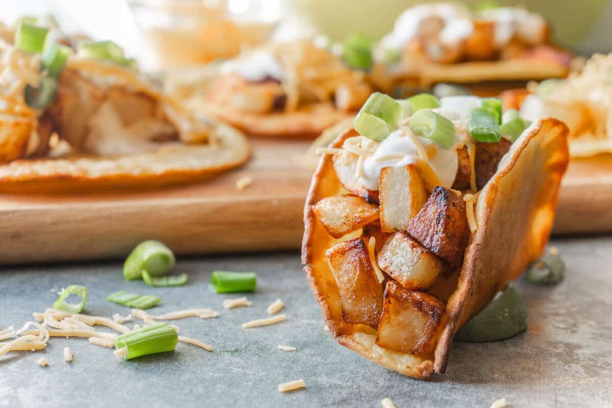 otato taco in crispy corn tortilla topped with sour cream, cheese, and green onion with more tacos on a wooden cutting board in the background.