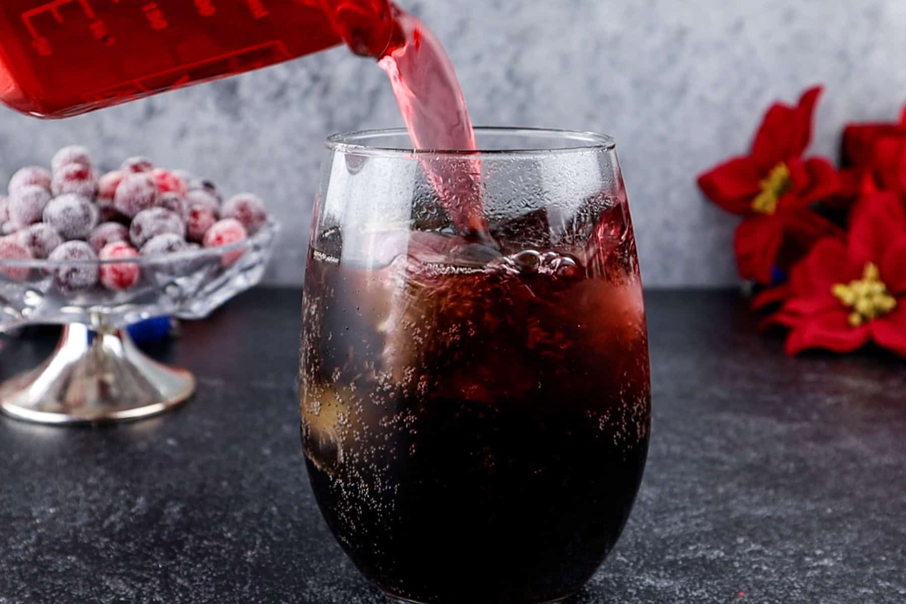 Adding cranberry juice to a glass with festive decorations in the background.