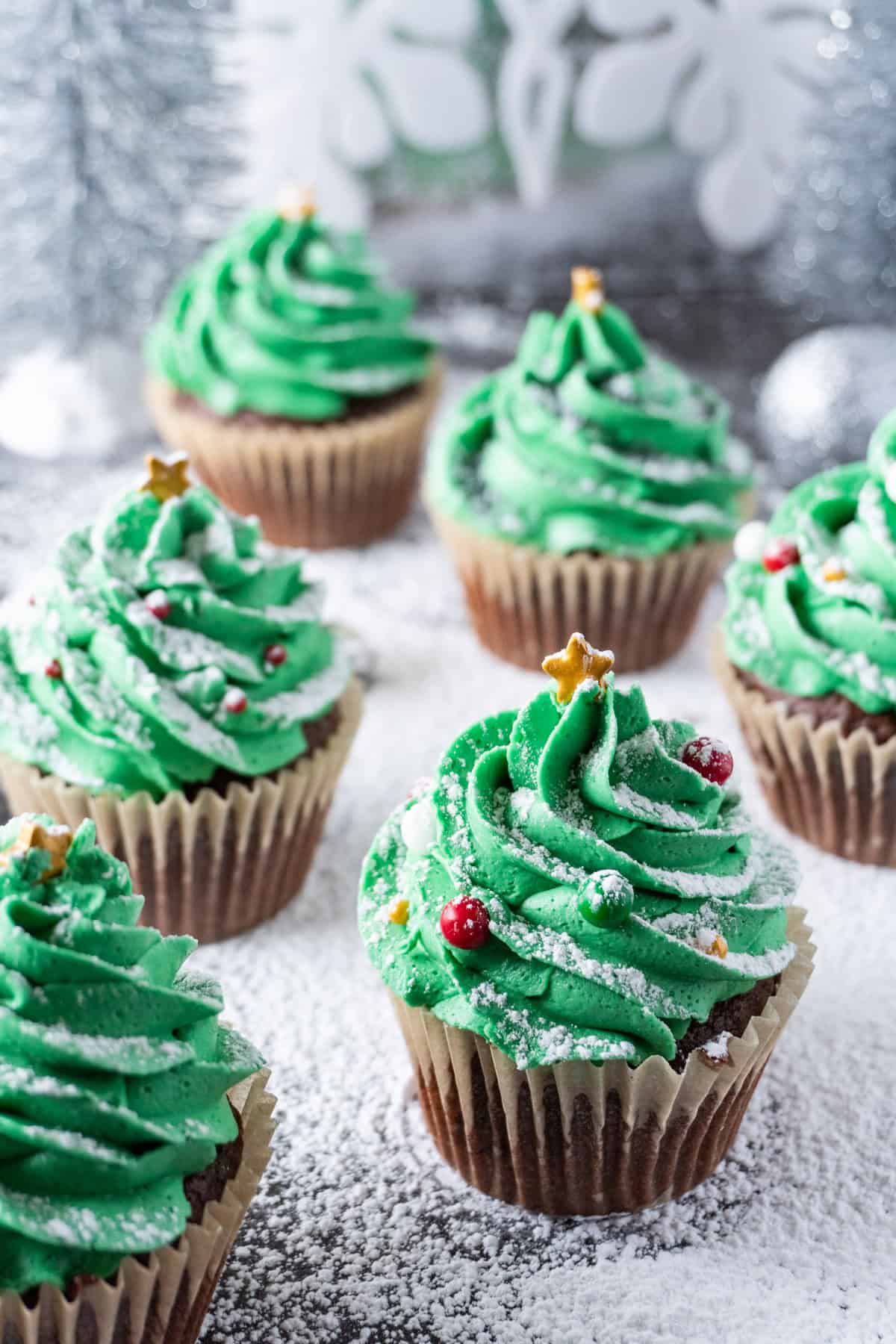 Christmas Tree Cupcakes with a powdered sugar sprinkled over them as snow and silver Christmas decorations in the background.