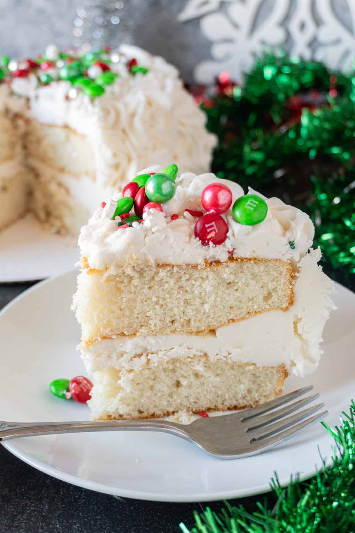 This easy Holiday Cake is beautiful, tasty, and surprisingly simple! #cake #christmascake #christmas #party #atablefullofjoy #easy #ideas #fancy #beautiful #simple #mix #decorating #recipes Slice of holiday cake on white plate with rest of cake in backround.