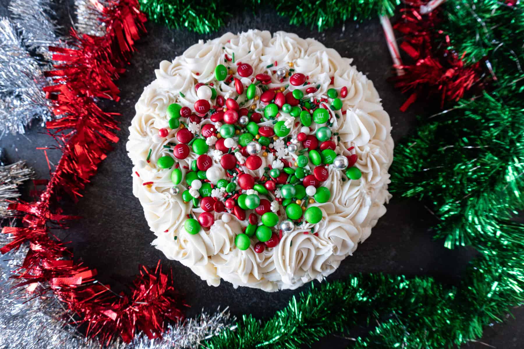 This easy Holiday Cake is beautiful, tasty, and surprisingly simple! #cake #christmascake #christmas #party #atablefullofjoy #easy #ideas #fancy #beautiful #simple #mix #decorating #recipes Shoiwng the top of the holiday cake, covered in buttercream swills, rend and green m&ms, and sprinkles.