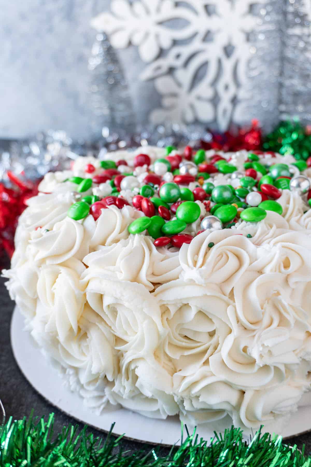 This easy Holiday Cake is beautiful, tasty, and surprisingly simple! #cake #christmascake #christmas #party #atablefullofjoy #easy #ideas #fancy #beautiful #simple #mix #decorating #recipes Easy holiday cake with piped on swirls and red and green decorations on top. Christmas decor in beackground.