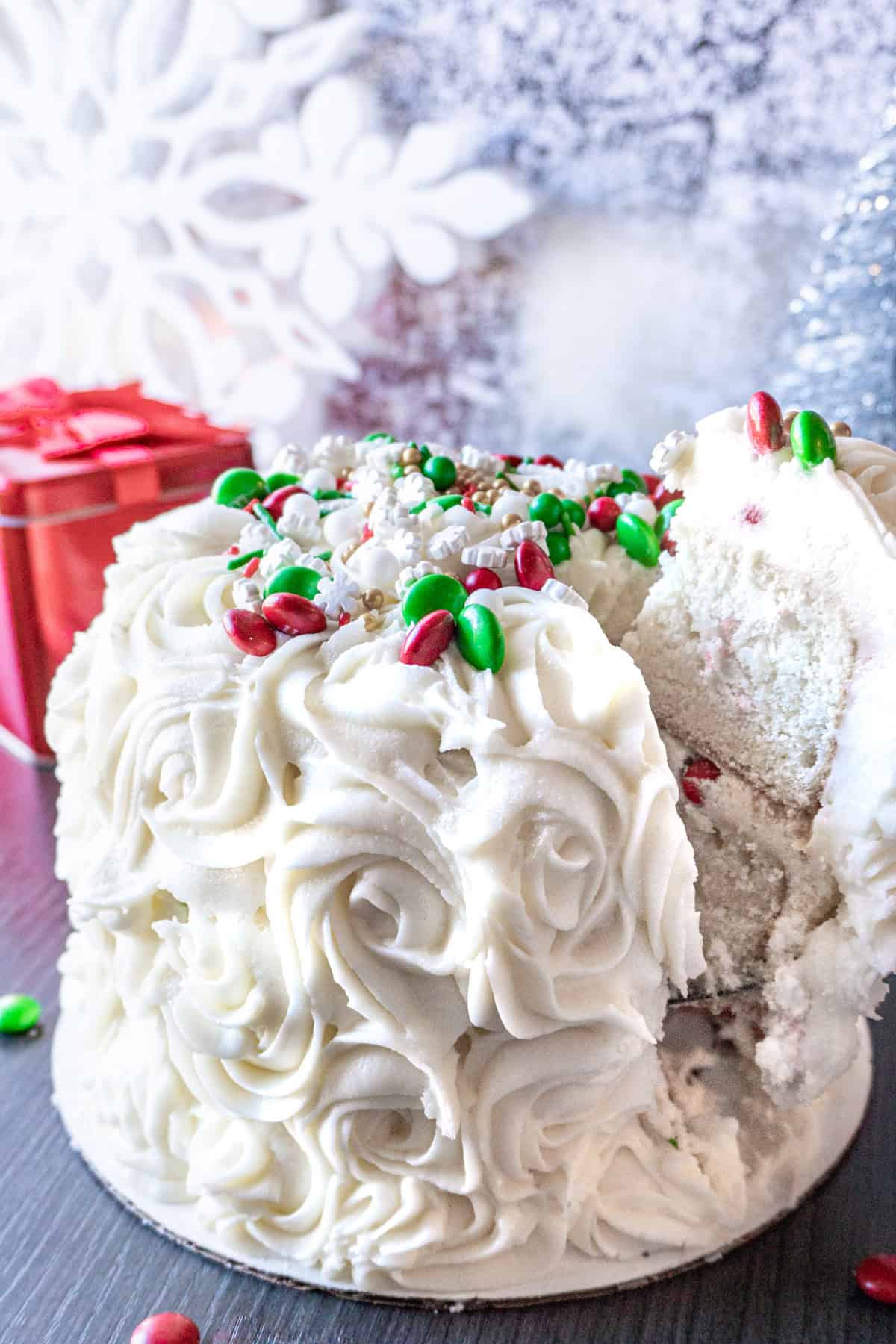 This easy Holiday Cake is beautiful, tasty, and surprisingly simple! #cake #christmascake #christmas #party #atablefullofjoy #easy #ideas #fancy #beautiful #simple #mix #decorating #recipes Take a slice of Holiday Cake decorated with white frosted rose swirls, M&Ms, and sprinkles, Christmas decorations in the background.