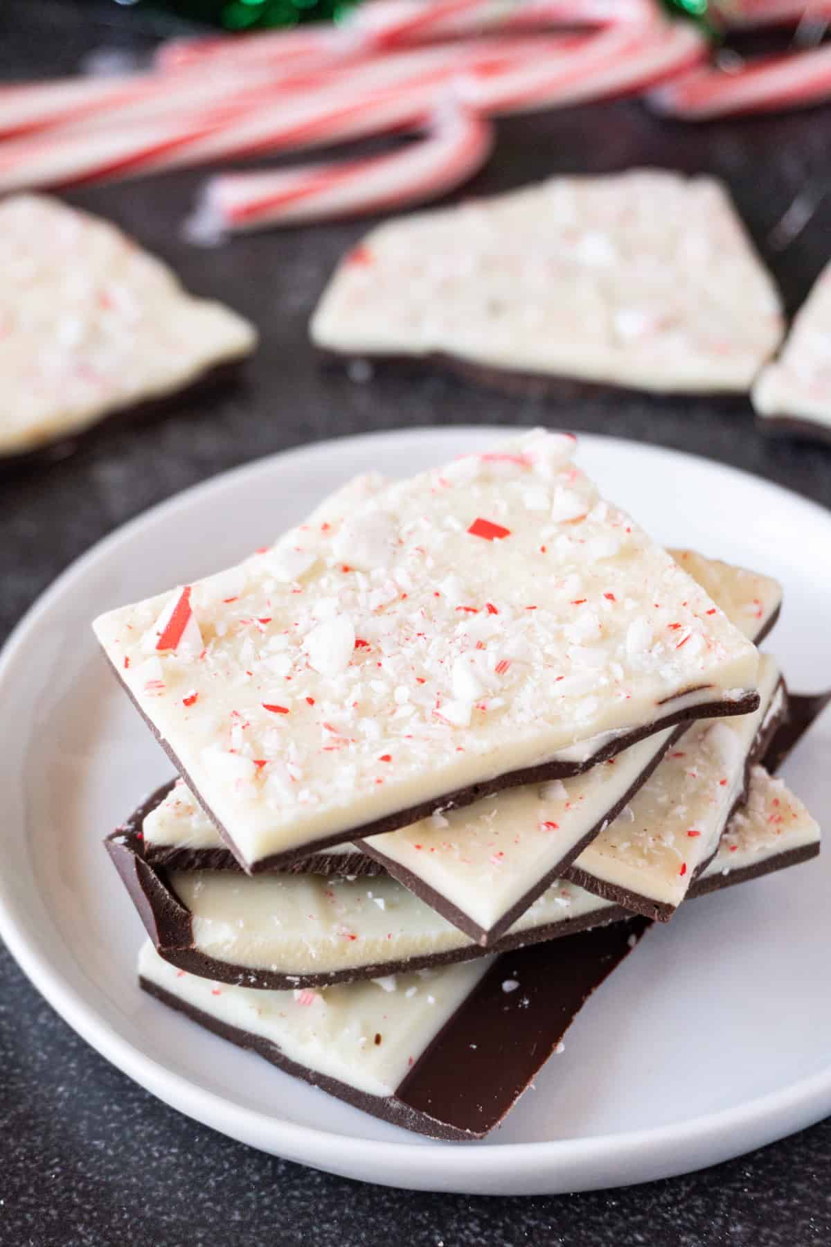Peppermint bark stacked on a white plate with more peppermint bark and candy canes in the background.