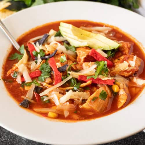 chicken tortilla soup in white bowl garnished with avocado, chips, green onions, featured image.