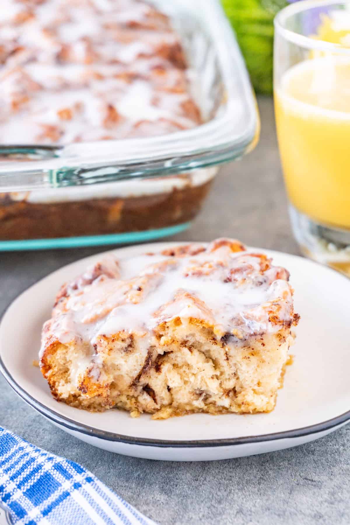 Slice of Cinnamon Roll French Toast on a white plate with glass of orange juice and casserole dish in background.