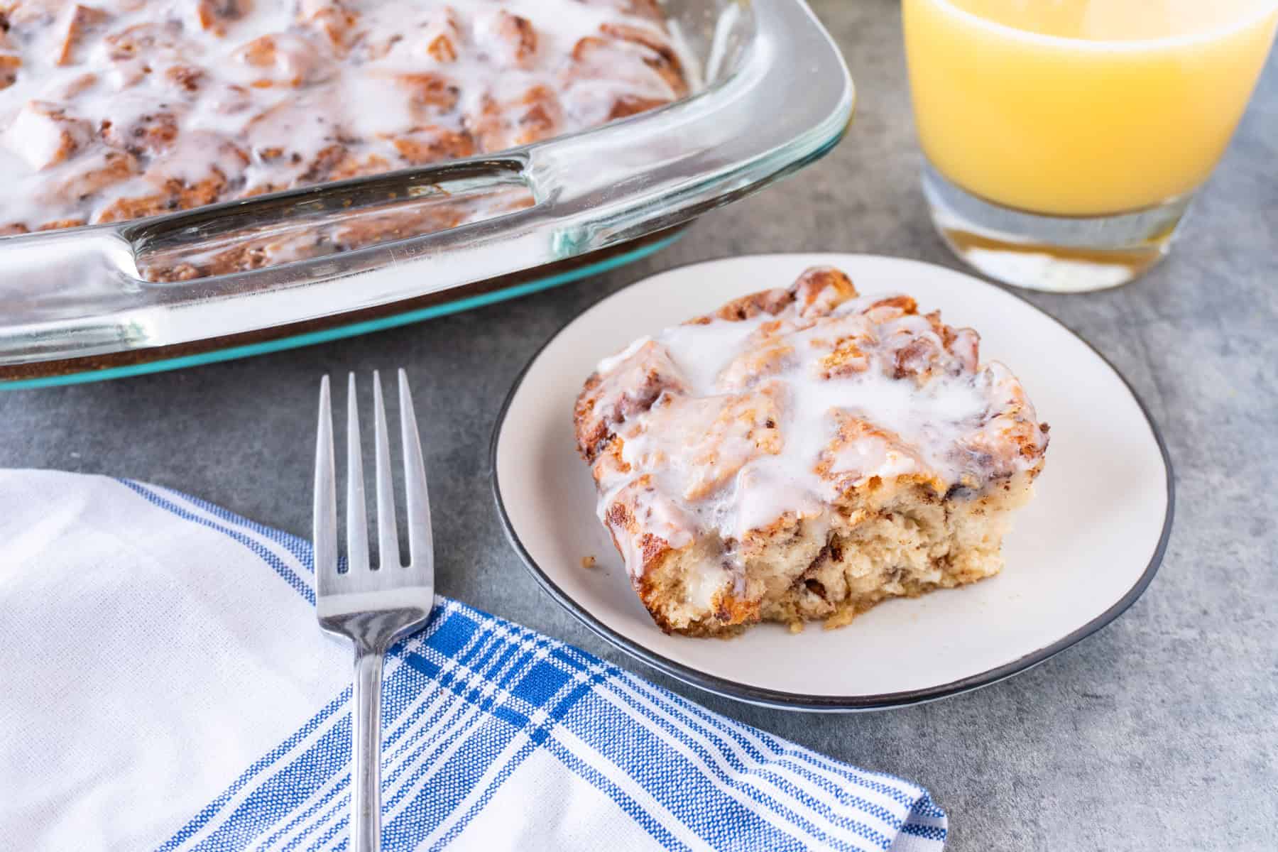 Slice of Cinnamon Roll Casserole on a white plate next to a fork and blue and white napkin.
