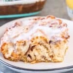Slice of Cinnamon Roll French Toast on a white plate next to blue and white napkin and glass of orange juice, pinterest image.