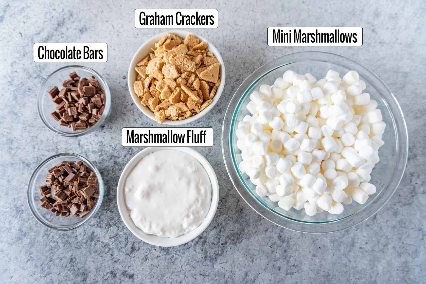 ingredients in bowls: Chocolate pieces, crushed graham crackers, marshmallow fluff, and mini marshmallows