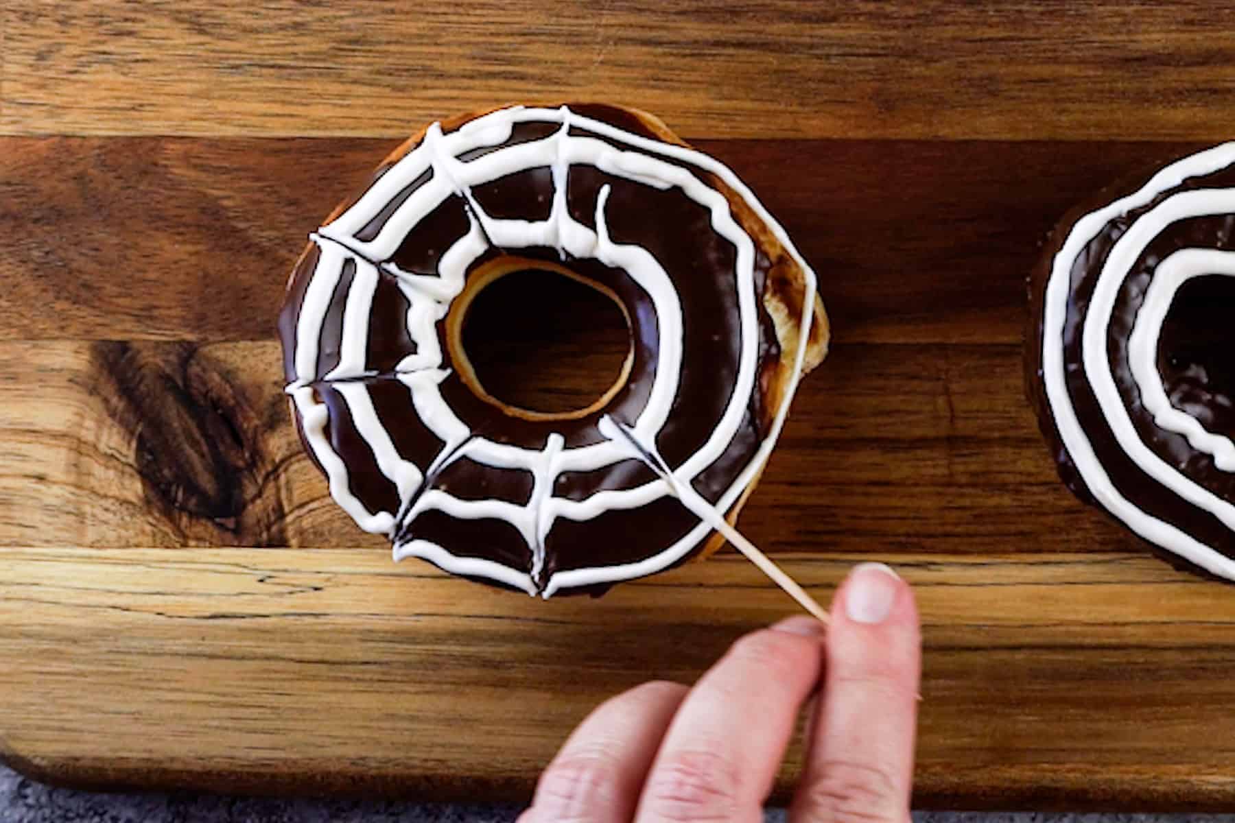 dragging a toothpick through the concentric white circle to make lines and form the web for the spiderweb donuts.