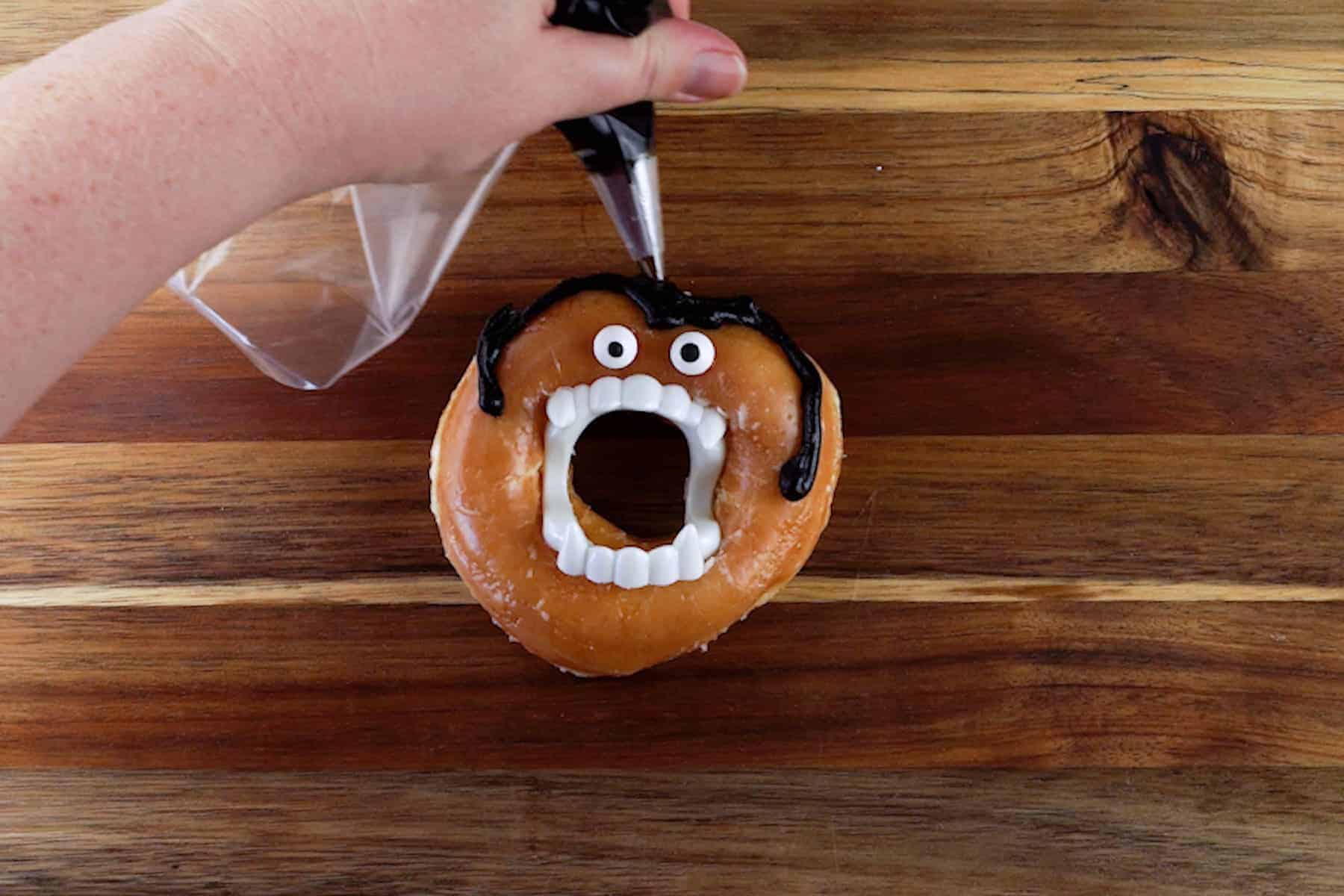 piping black frosting as hair to a glazed vampire donut.