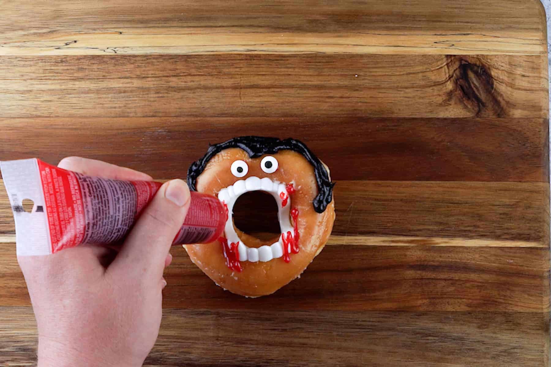 adding red gel icing as blood around plastic vampire fangs on glazed donut.