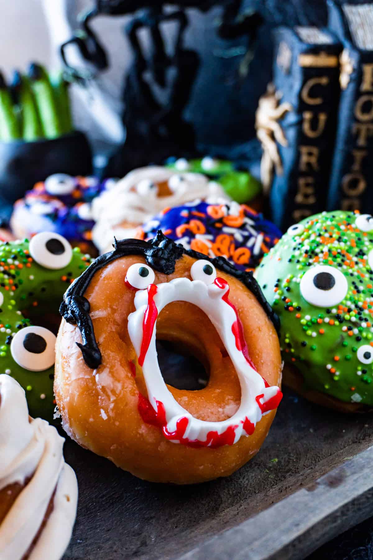 Dracula decorated glazed donut surrounded by other Halloween decorated donuts on a wooden dish.