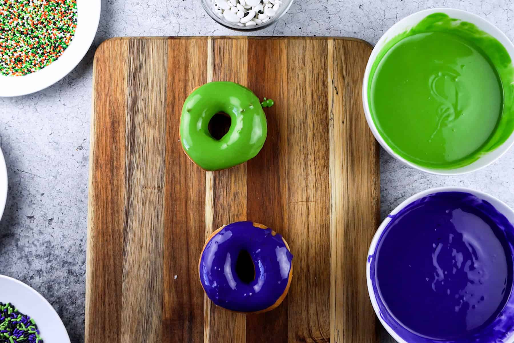 glazed donuts dipped in green and purple candy melts on a wooden cutting board.