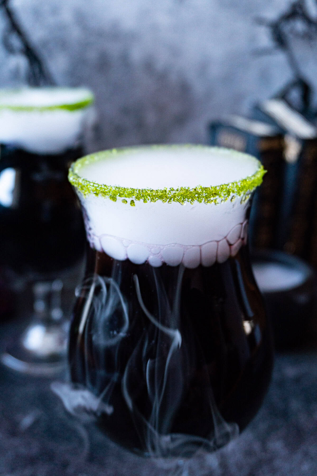 black cauldron cocktail in a clear glass cup with a green sugared rim and with dry ice "smoke" spilling out, and Halloween decorations in the background.