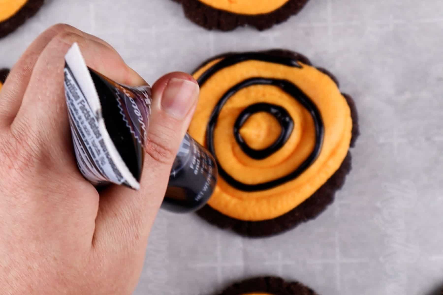 Piping concentric circles of black gel onto a cookie frosted with orange frosting.