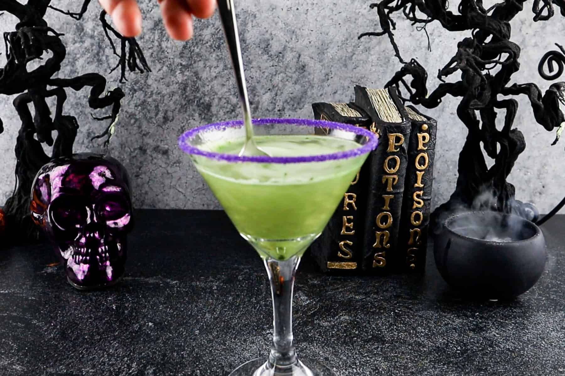 stirring green and gold luster dust into the cocktail, Halloween decorations in the background.