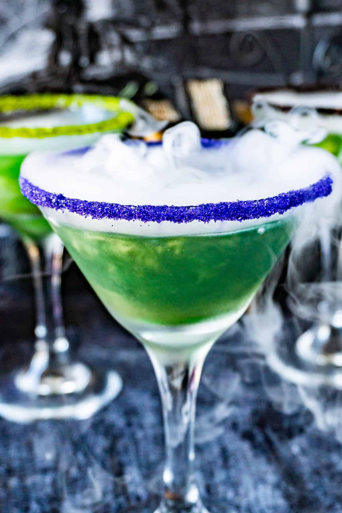 witches brew cocktail in a martini glass with purple sanding sugar on the rim and dry ice "smoke" spilling out of the cup.