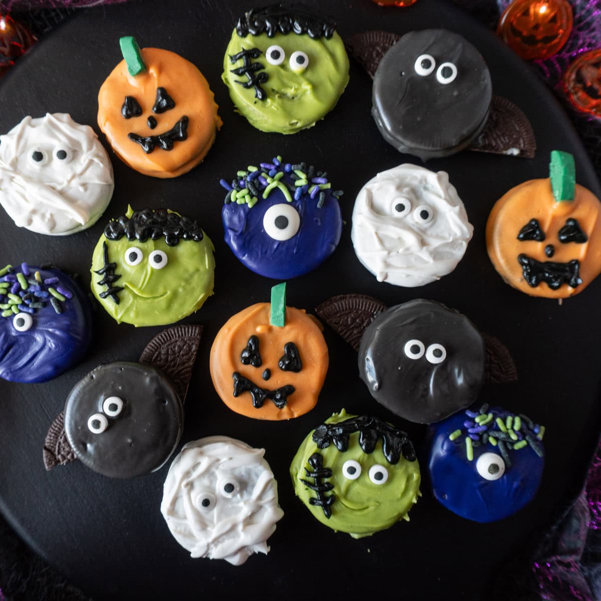 Halloween Oreos all decorated as different Halloween Themes on a black background surrounded by pumpkin lights and purple ribbon, featured image.