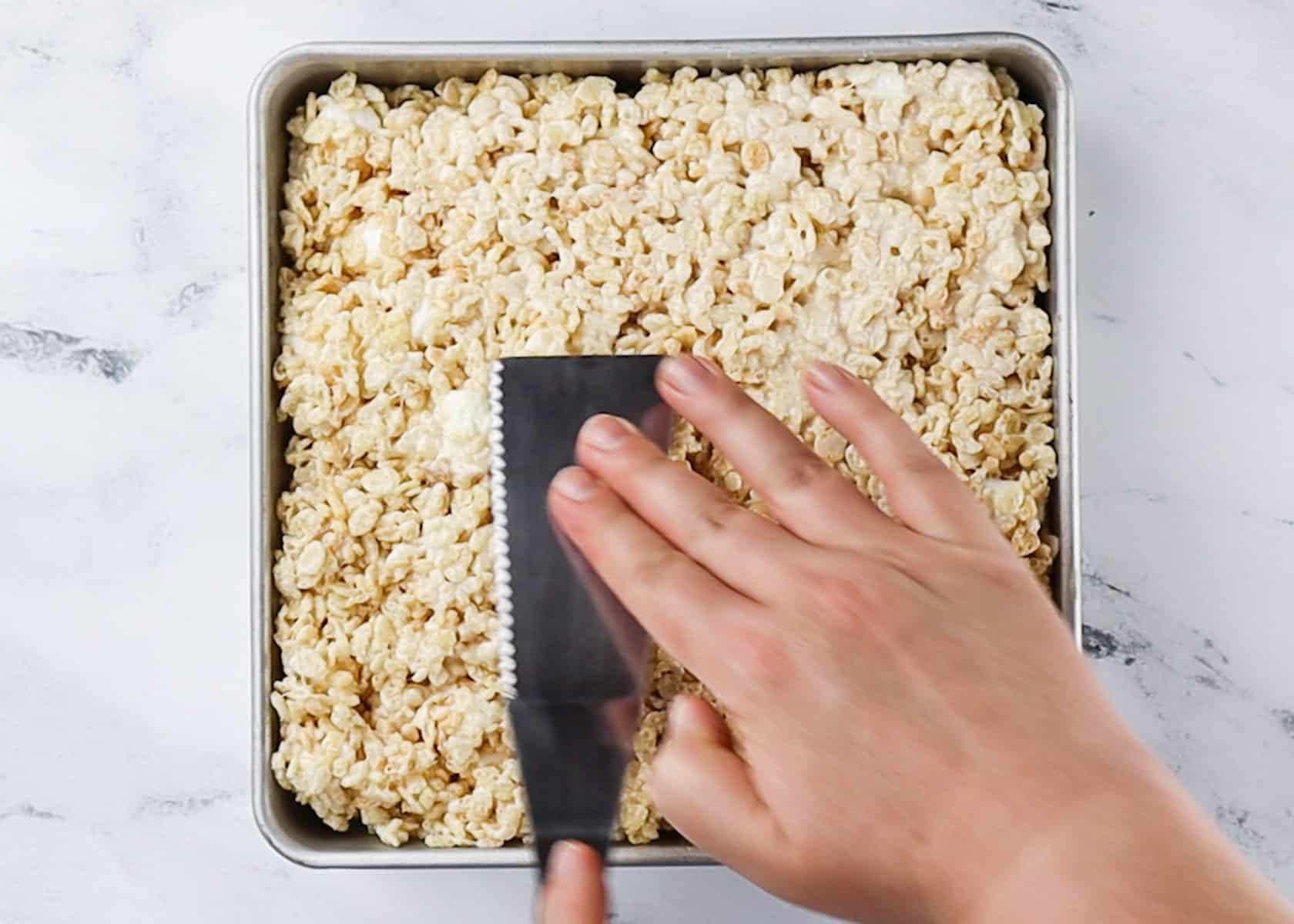 Patting rice krrispies treats down into a metal square baking pan.