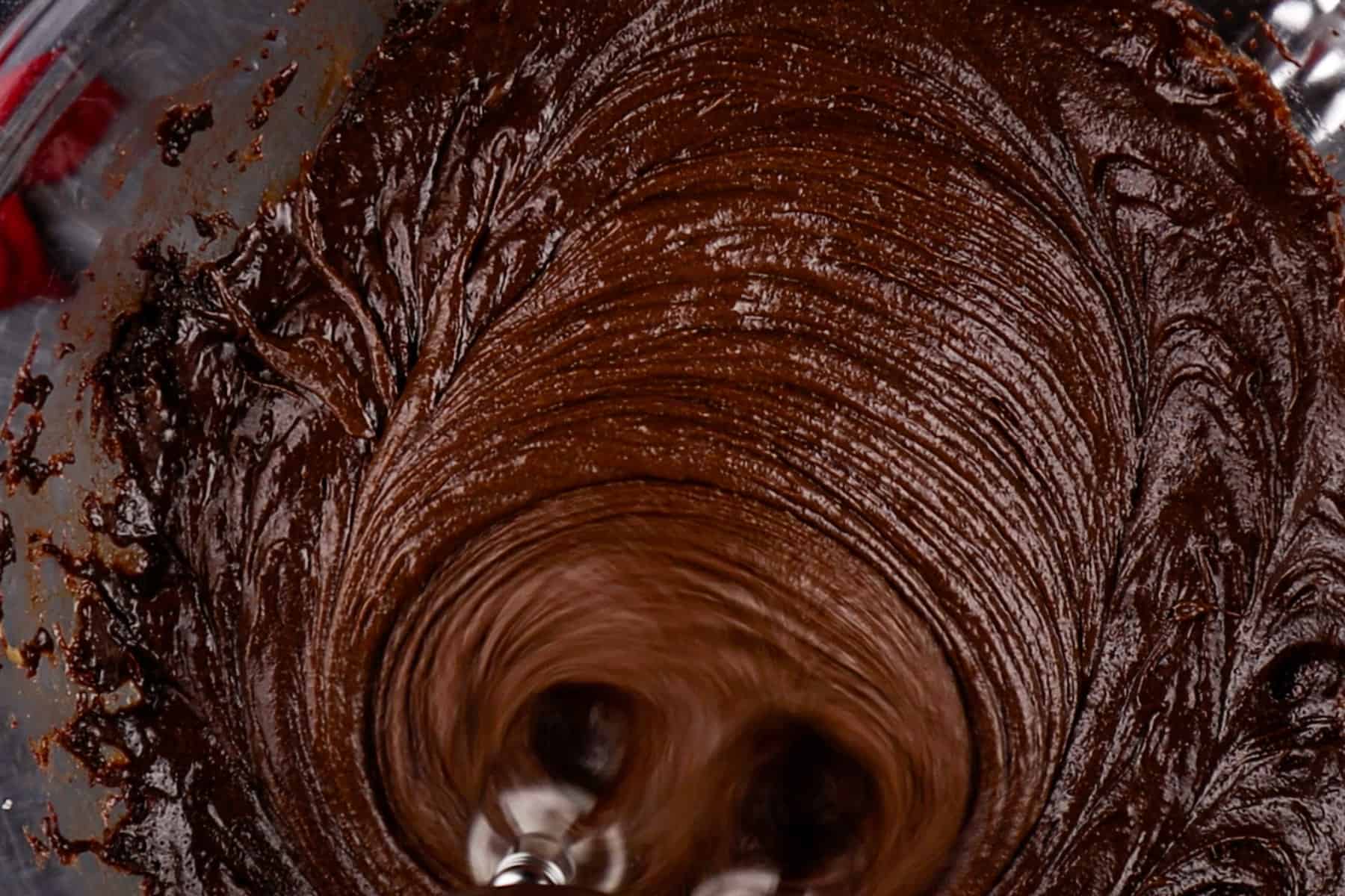 Brownie batter is being mixed with a hand mixer in a glass bowl.