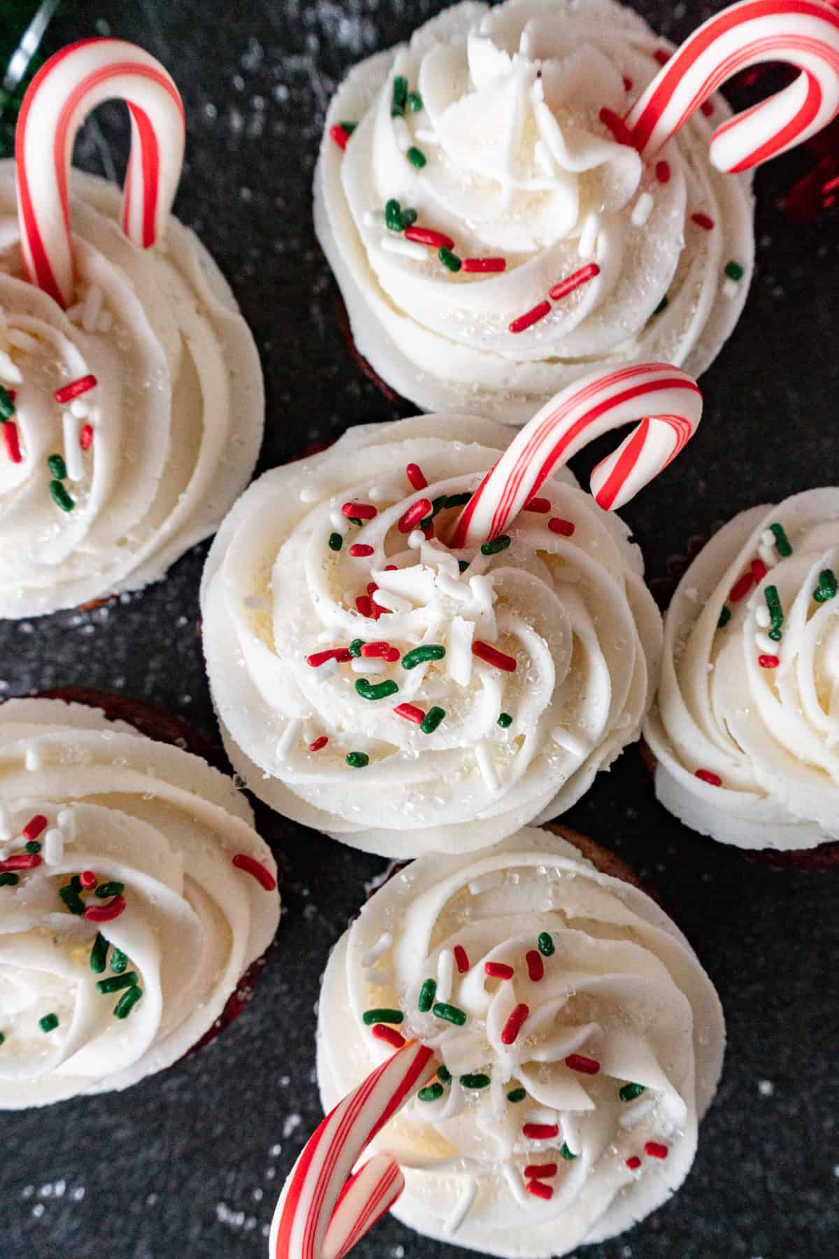 Candy cane cupcakes close together, image of the tops of the cupcakes, showing the white frosting, sprinkles and candy canes.