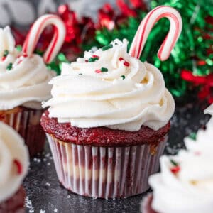 Candy cane cupcakes, with peppermint frosting, a candy cane stuck in the frosting, and red and green sprinkles on top. Christmas decorations in background, featured image.