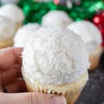 holding a snowball cupcake with sparkly sanding sugar on top, with more cupcakes in background.