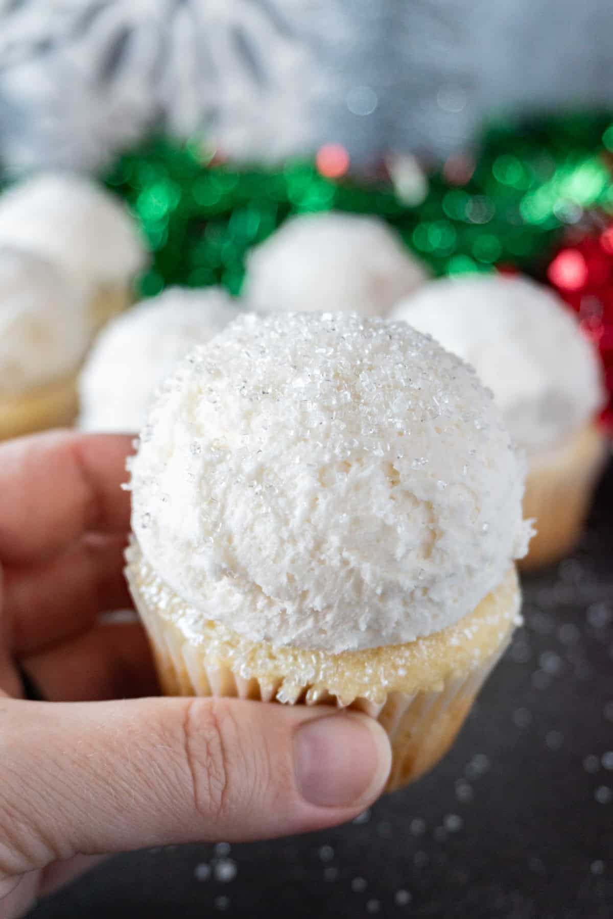 holding a snowball cupcake with sparkly sanding sugar on top, with more cupcakes in background.