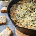 Hot artichoke dip in a cast iron pan on wooden trivet with pieces of bread scattered around.