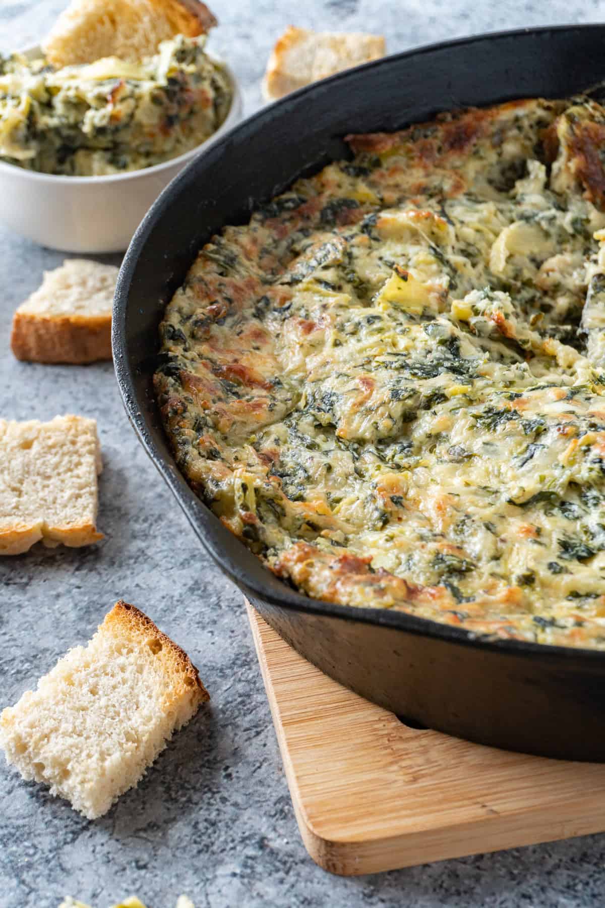 Hot artichoke dip in a cast iron pan on wooden trivet with pieces of bread scattered around.