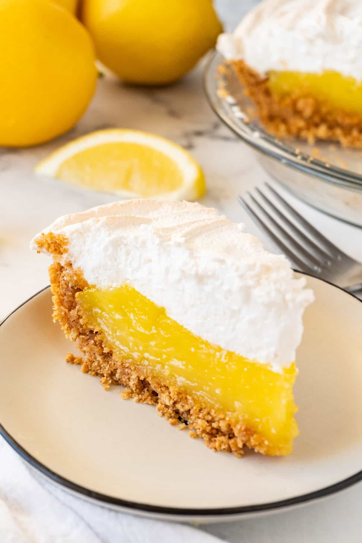 slice of lemon meringue pie on white plate with lemons in background.