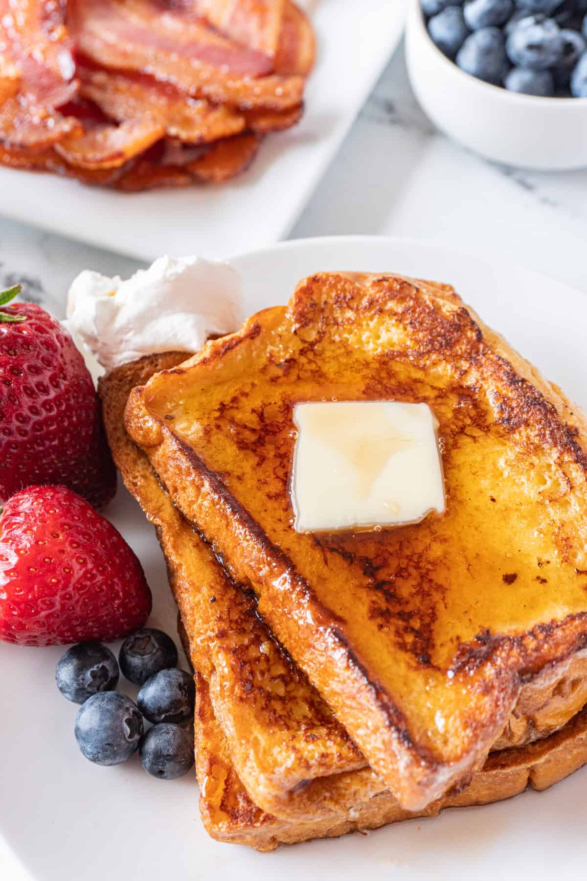 french toast with butter and berries and syrup on a plate with bacon in background