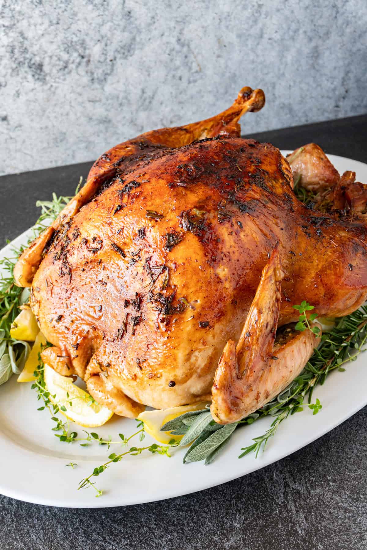 Herb butter turkey with golden skin on a white platter surrounded by fresh herbs and lemon slices.